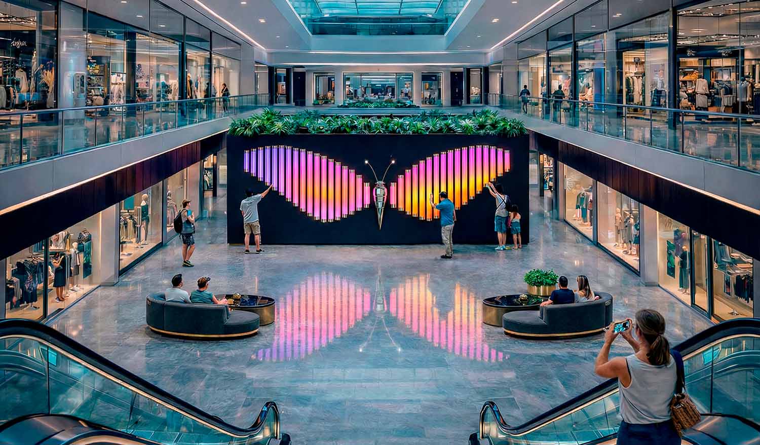 Интерактивная световая инсталляция «Бабочка» в центральном атриуме торгового центра: крупные крылья из вертикальных LED-труб формируют розово-оранжево-фиолетовый градиент и отражаются в глянцевом полу; посетители взаимодействуют со скульптурой, меняя свет при прикосновении.