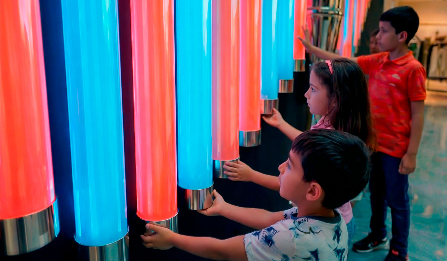 Крупный план интерактивной световой инсталляции: дети прикасаются к вертикальным LED-трубам бирюзового и розового цвета, активируя изменение света; глянцевые цилиндры с металлическими креплениями реагируют на прикосновение.