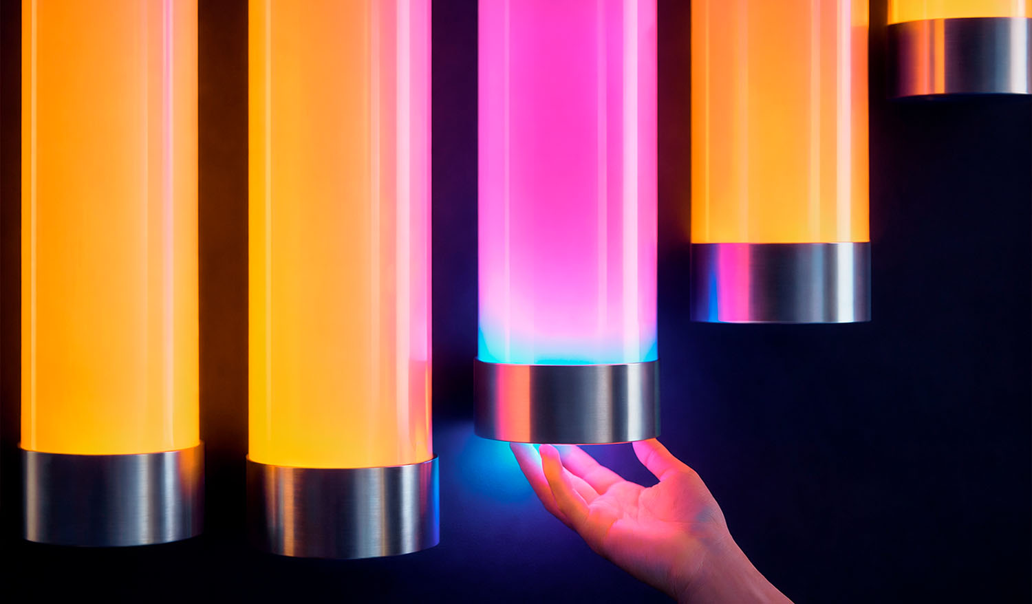 Деталь интерактивной световой инсталляции: рука подносится к вертикальной LED-трубе с градиентом розового и голубого цвета, активируя изменение света; глянцевый цилиндр с металлическим основанием подсвечивается в ответ на прикосновение.