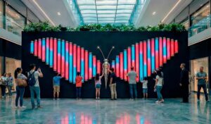 Interactive light installation shaped as a neon butterfly in a shopping mall atrium: vertical pink and cyan LED tubes respond to visitors’ touch; a metallic central body with antennas anchors the composition as people engage with the artwork.