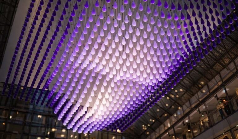 A kinetic installation of hundreds of teardrop-shaped units illuminated in purple, forming a three-dimensional face beneath the glass dome of a modern atrium.
