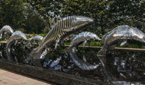 Металлические скульптуры рыб в проекте «Водный мир» с полосатой и перфорированной текстурой, отражающиеся в тёмной глади воды парка.