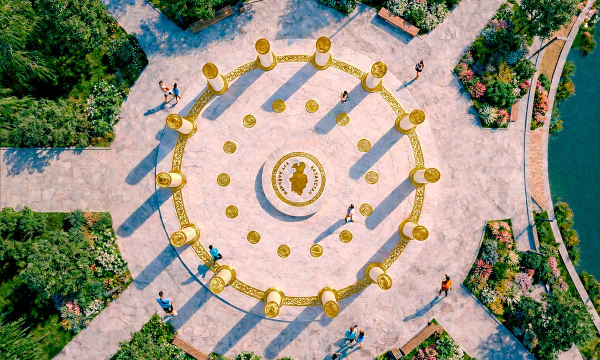 Вид сверху на инсталляцию «Богатство Нации» — 14 колонн образуют круг вокруг центральной платформы с картой Казахстана, золотые медальоны встроены в мраморную площадь; композиция расположена в озеленённом парке у воды, посетители гуляют по периметру.