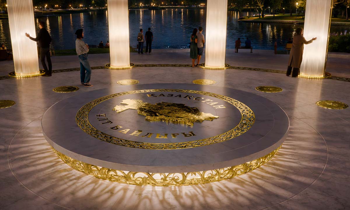 Центральная платформа инсталляции «Богатство Нации» с рельефной картой Казахстана и надписью «Қазақстан — Ұлт байлығы», окружённая светящимися колоннами у набережной; вечерняя подсветка создаёт золотые узоры на мраморной поверхности, посетители взаимодействуют с пространством.