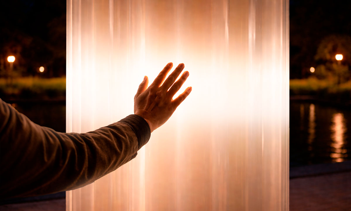 Рука человека касается светящейся колонны интерактивной инсталляции ночью; тёплый свет мягко подсвечивает ладонь, на фоне видна набережная и отражения огней в воде.