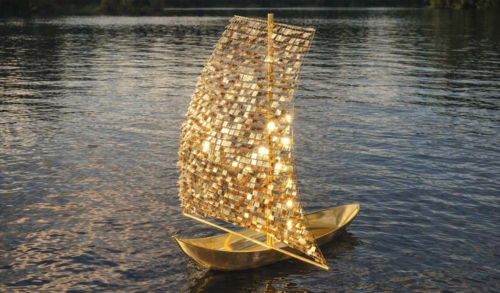 Художественная инсталляция в виде золотого паруса на воде — скульптура с мозаичной отражающей поверхностью и металлическим корпусом лодки, мягко сияющая в естественном свете на фоне спокойной глади озера.