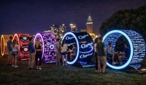 Интерактивная инсталляция «Городские порталы» — ряд светящихся круглых капсул с неоновой подсветкой и брендированной графикой, установленных на газоне в вечернем городе; внутри посетители сидят за столиками и общаются, а яркие цветовые акценты создают эффектный медиа-образ на фоне ночного города.