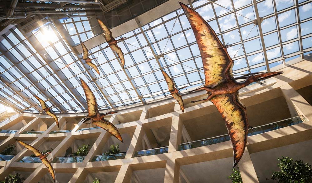 Кинетическая инсталляция с крупными фигурами птеродактелей под стеклянной крышей современного атриума, солнечный свет проходит через прозрачный потолок и освещает многоуровневое пространство с балконами и растениями.