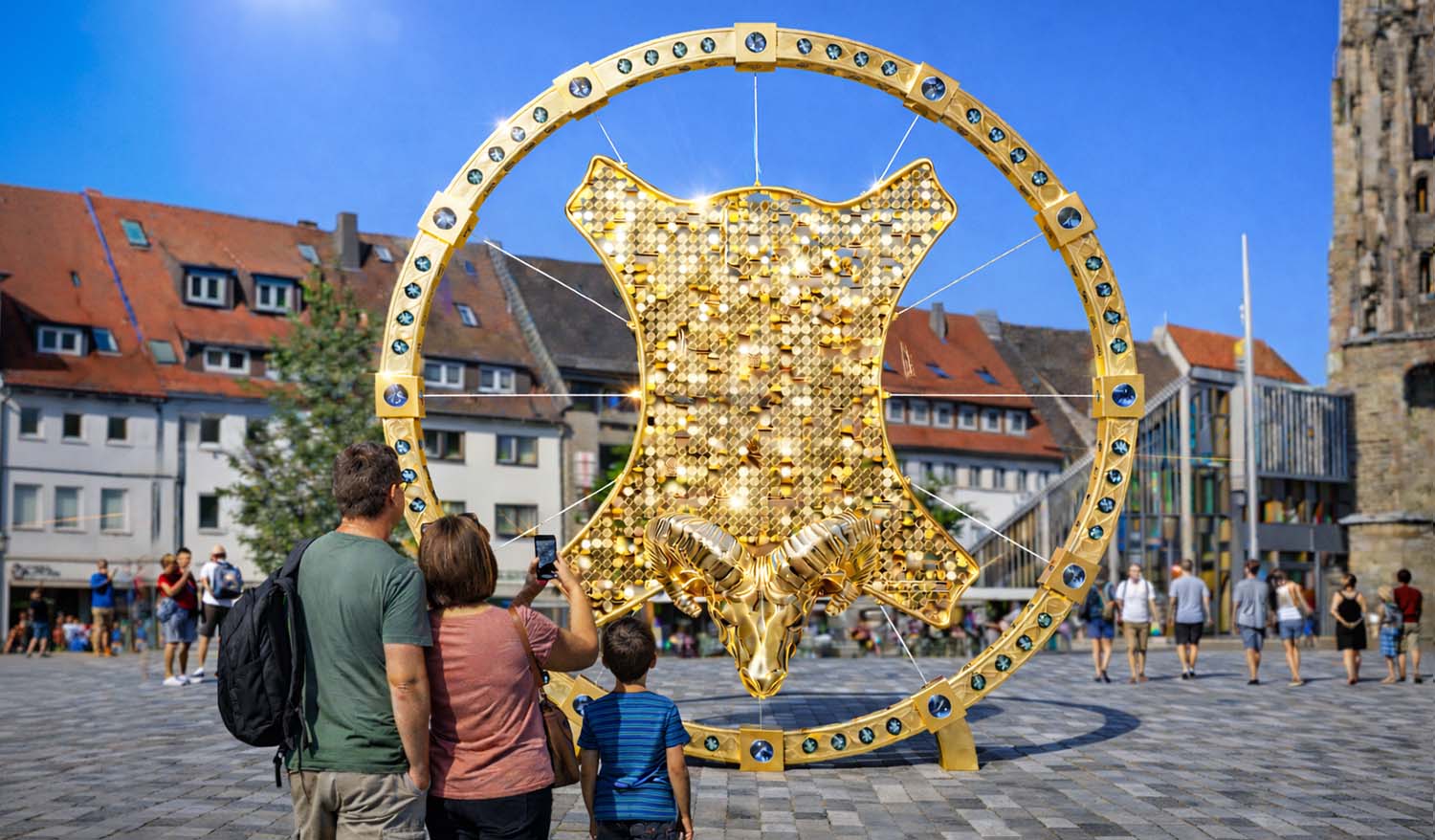 Кинетическая инсталляция «Золотое руно» на городской площади днём — золотая круглая рама с декоративными вставками, внутри которой мерцает панно из отражающих элементов и объемная голова барана в нижней части; семья рассматривает арт-объект.