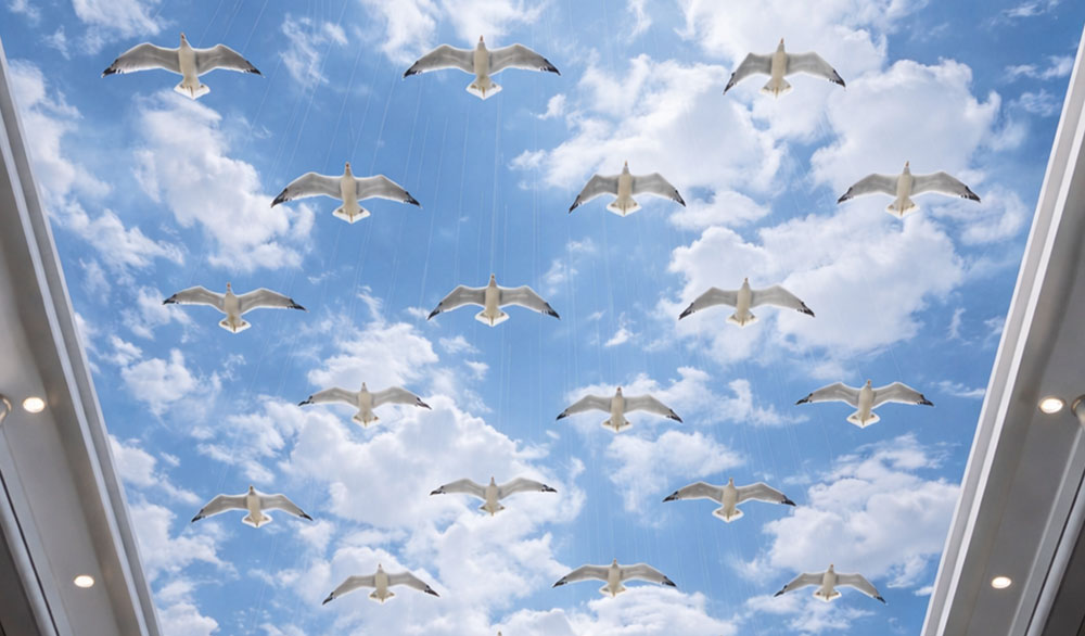 Кинетическая инсталляция «Чайки» от Skyform Studio — ряды белых птиц с расправленными крыльями подвешены на почти незаметных тросах под стеклянной крышей, на фоне яркого голубого неба с облаками.
