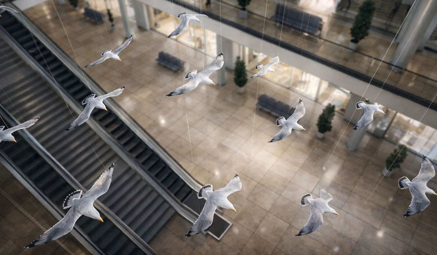 Фрагмент кинетической инсталляции «Чайки» — белые птицы с детализированными крыльями и жёлтыми клювами подвешены на тонких тросах над зоной эскалаторов; вид сверху подчёркивает разную высоту размещения элементов и ощущение плавного движения стаи в пространстве светлого атриума.