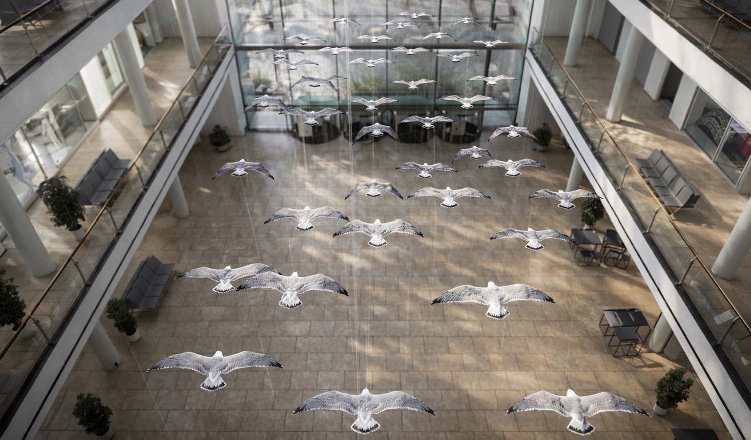 Вид с верхнего уровня атриума на кинетическую инсталляцию «Чайки» — десятки белых фигур птиц подвешены на тонких тросах и расположены на разных высотах, солнечный свет из стеклянного фасада мягко подсвечивает крылья.
