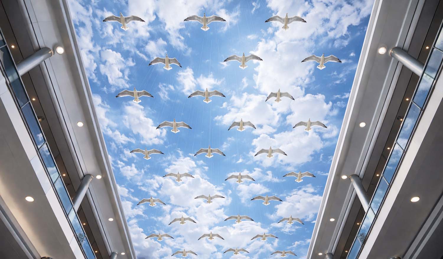 Вид снизу на кинетическую инсталляцию «Чайки» — десятки белых фигур птиц с расправленными крыльями подвешены под крышей атриума, выстраиваясь в ритмичный узор на фоне яркого небесного принта с облаками, создавая иллюзию стаи, летящей в открытом небе.