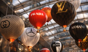 Кинетическая инсталляция «Shopping Vortex» — подвесная композиция из брендированных воздушных шаров Prada, Louis Vuitton, Gucci, Armani, Boss и Mercedes под стеклянным куполом, с тёплой подсветкой, создающей эффект роскошного вихря покупок.