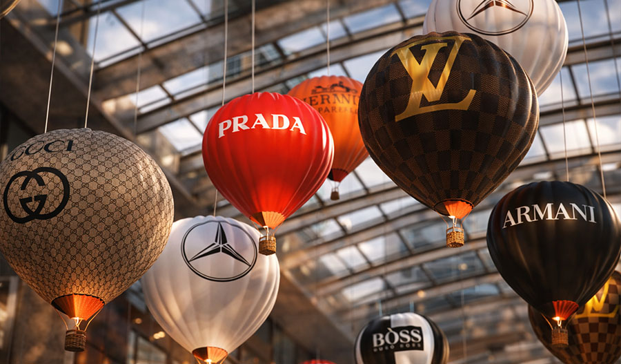 The kinetic installation "Shopping Vortex" is a suspended composition of branded balloons from Prada, Louis Vuitton, Gucci, Armani, Boss, and Mercedes under a glass dome, with warm lighting creating the effect of a luxurious shopping whirlwind.
