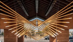 Кинетическая инсталляция «Крылья долины» — масштабная композиция из золотистых труб, формирующих симметричные крылья в пространстве под сводчатой крышей; динамичная геометрия и тёплая подсветка создают эффект парящего объёма и архитектурный акцент входной зоны.