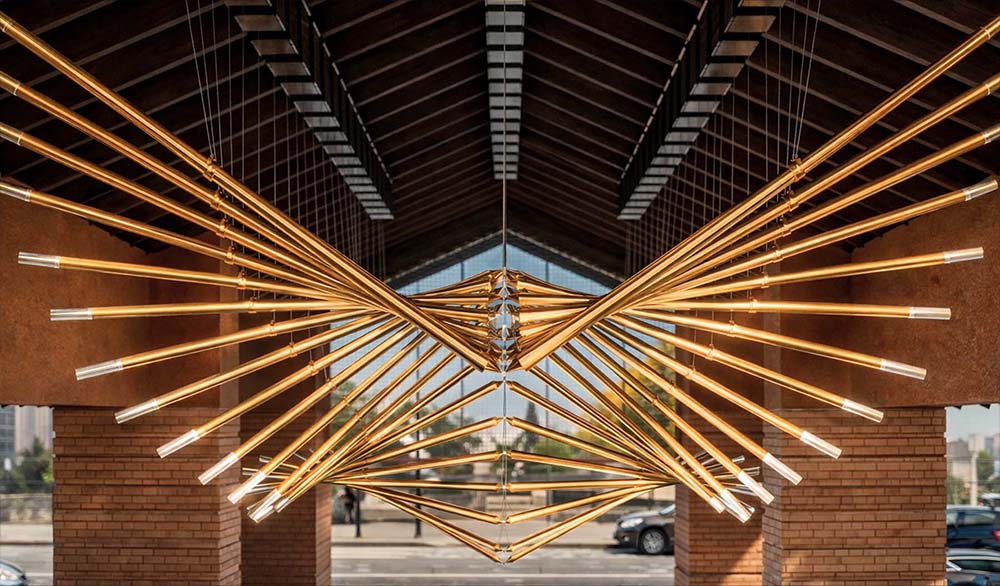 Кинетическая инсталляция «Крылья долины» — масштабная композиция из золотистых труб, формирующих симметричные крылья в пространстве под сводчатой крышей; динамичная геометрия и тёплая подсветка создают эффект парящего объёма и архитектурный акцент входной зоны.