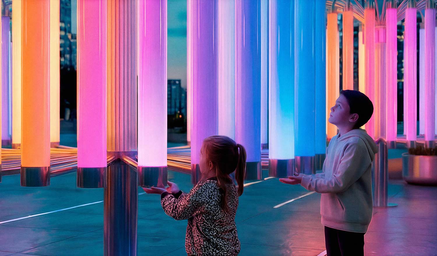 Интерактивная инсталляция «Пульс Света»: дети подносят руки к светящимся поликарбонатным трубам, активируя пульсирующие цветовые переходы (розовый, голубой, оранжевый) и создавая живой светозвуковой отклик в вечернем городском пространстве