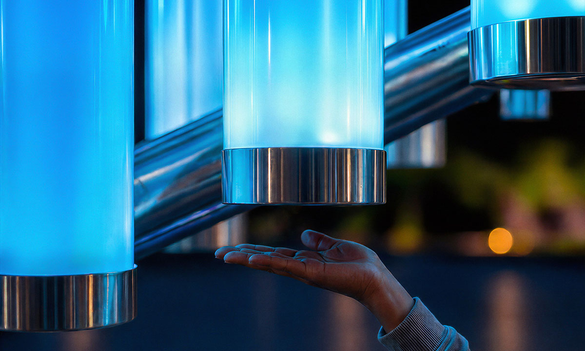 Close-up of the interactive installation “Magic Streams” — an illuminated cylinder made of matte polycarbonate with metal elements, responding to the approach of a hand; the soft blue glow intensifies upon interaction, creating a responsive light effect.