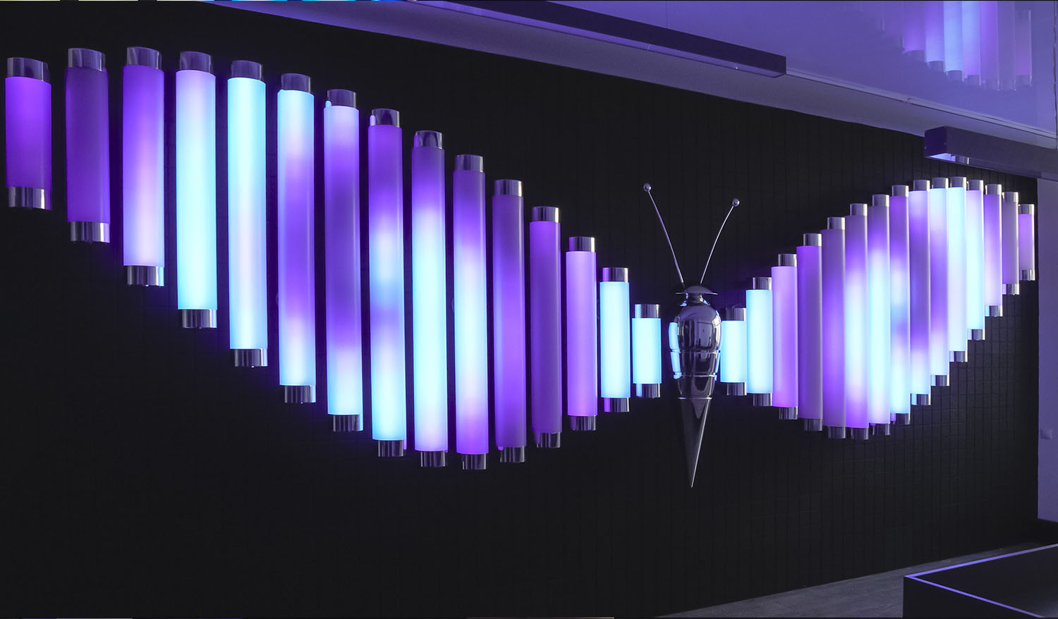 Interactive Butterfly light installation with wings made of glowing violet and cyan tubes and a metallic butterfly sculpture at the center.
