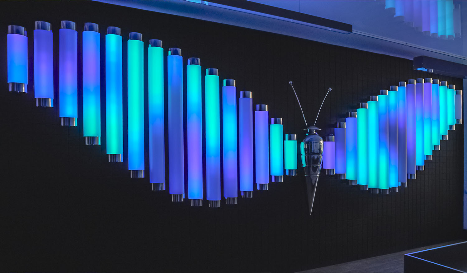 Interactive Butterfly light installation with wings made of glowing blue, turquoise, and violet tubes and a metallic butterfly sculpture at the center.