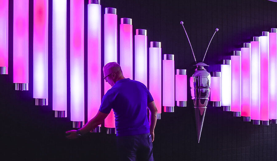 Odwiedzający wchodzi w interakcję z instalacją „Butterfly”, złożoną z pionowych tub świetlnych świecących na różowo i biało.